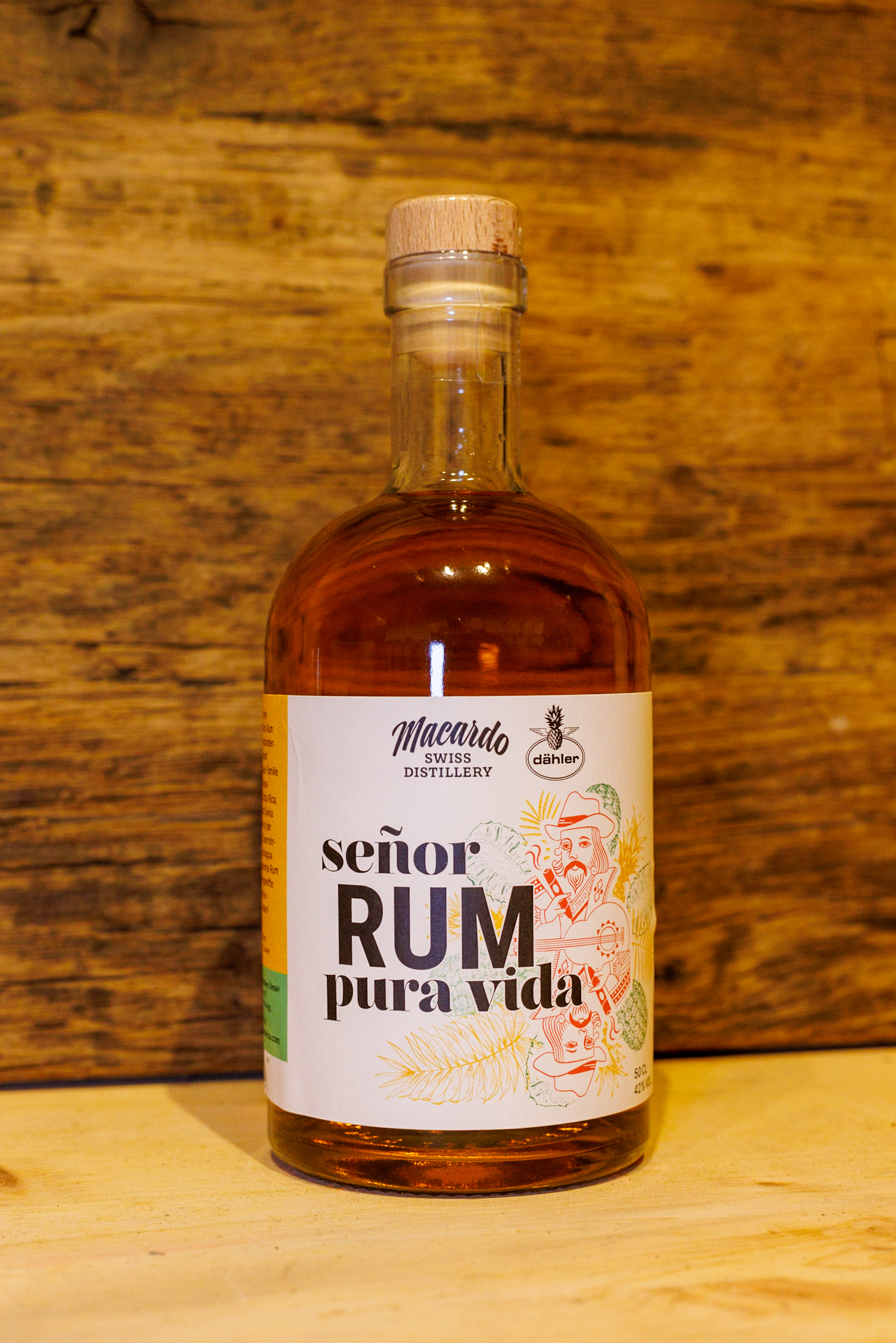 Eine Rum Flasche der Macardo Swiss Distillery steht vor einer Holzwand. Das Etikett des "Señor Rum Pura Vida" zieren Illustrationen eines Mannes mit Gitarre und tropische Motive wie Ananas und Palmenblätter. Der Rum hat eine goldene Farbe.